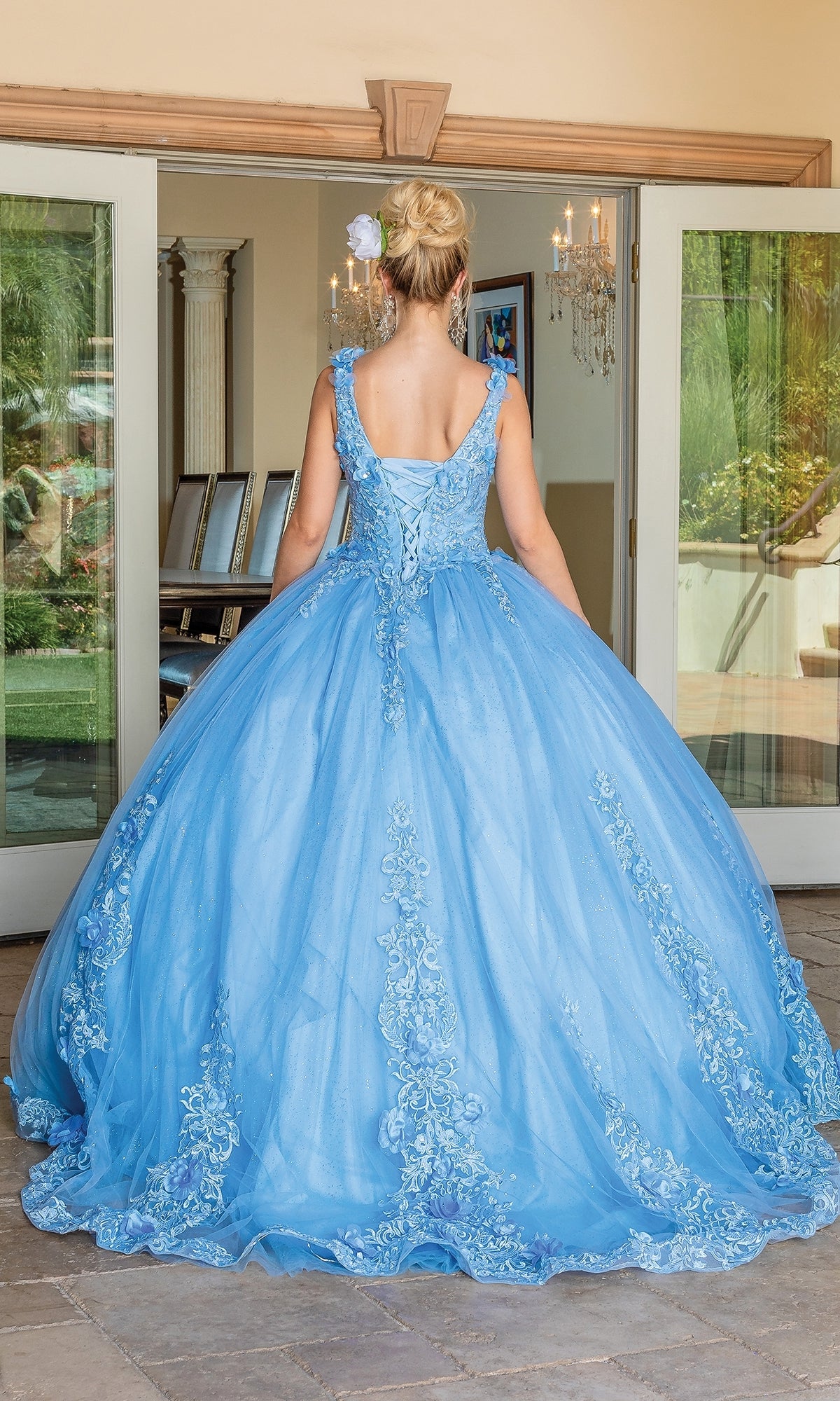 A woman with blonde hair in an updo wears the Quinceanera Dress 1678 By Dancing Queen, a floor-length light blue A-line ball gown with lace details and removable draped sleeves, standing facing glass doors from behind.